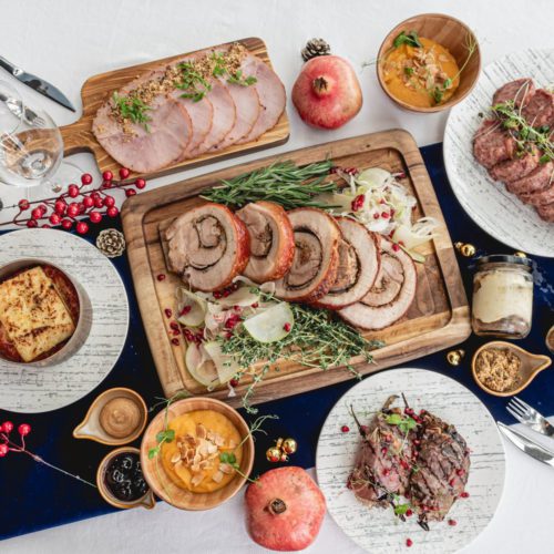 A table set with wooden boards and plates of food in Monti restaurant's Christmas Bundle available for delivery during Christmas 2020. Image depicts slices of roasted ham, thick slices of roulade, pork lasagna, oven baked wagyu with a sprinkling of red pomegranate seeds; and tiramisu in jars. 