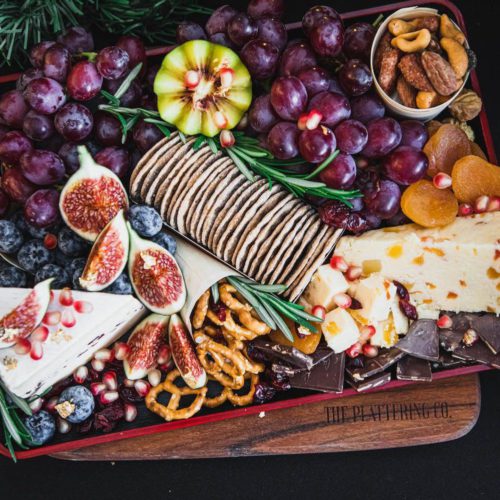 A cheese platter on a wooden serving board by The Plattering Co. has bunches of large purple grapes, slices of figs, blueberries, dried apricot, mixed nuts and dried fruit, pretzels and chocolate thins. Two wedges of special Christmas cheeses. Garnished with pomegranate seeds and sprigs of rosemary
