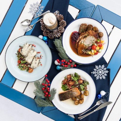 A blue and white wooden table evoking Grecian colours. Three plates of food from Greek restaurant's Zorba Christmas Bundle for 2 is shown. There is a choice of Roasted Turkey with Rice or the Grilled Salmon for the mains. Dessert is sweet Baklava pastry. A Christmas drink in a glass mug is shown, surrounded by pine cones.