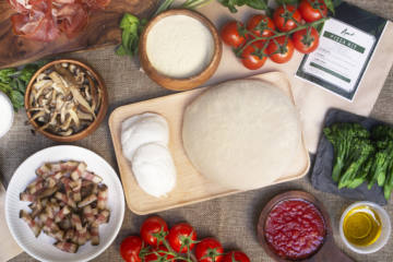 Picture of Italiamo Pizza kit from Amo restaurant, with slow-fermented dough, buffalo mozzarella, extra virgin olive oil, sauteed mushroom, vine tomatoes, guanciale and flour on a brown place mat, and broccolini served on a slate