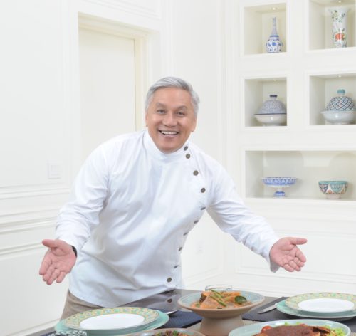 Picture of Datuk Chef Wan smiling in chef's whites, arms outstretched over  pretty plates on a lacquered table