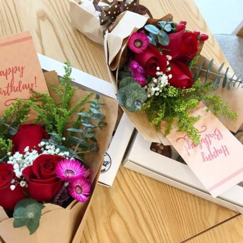 Complete your Valentine's Day dinner with a box of 6 assorted cupcakes with two petite bouquets of 3 roses and A6 happy birthday card from Baker V on a wooden table
